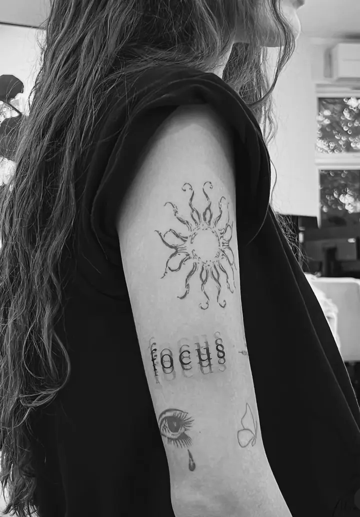 Sol, ordet «focus» og øye på overarmen, blackwork av Abas