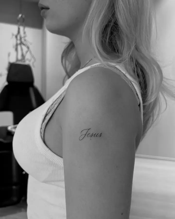 Ordet «Jesus» i skrift på overarmen, fineline av Abas