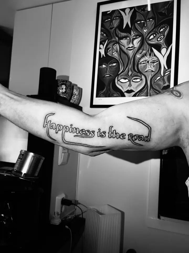 Teksten «Happiness is the road» på underarmen, blackwork av Sandra